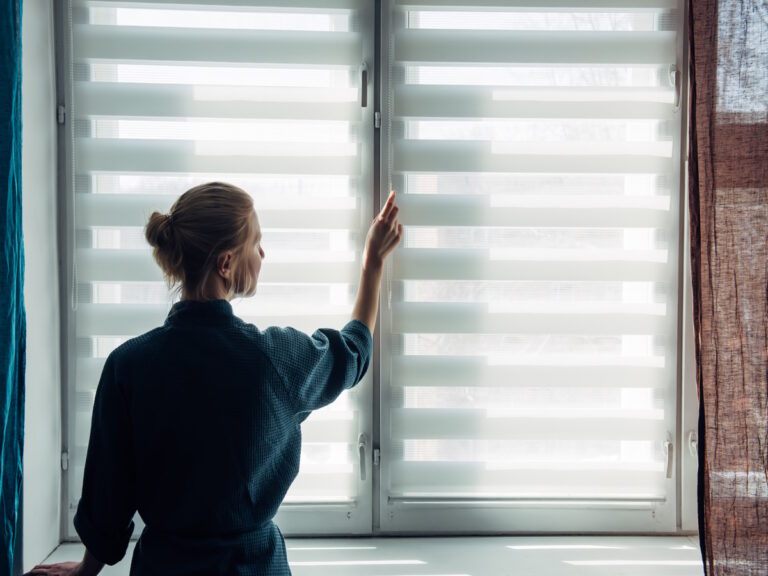 Woman's silhouette at window in the background light. Young woman in gown stands near window with blinds. Quarantine, self-isolation, stay home, self-preservation, coronavirus pandemic.